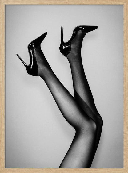 A black and white photograph of a woman's legs in the air, wearing sheer black stockings and shiny black stiletto high heels against a plain grey background. Poster