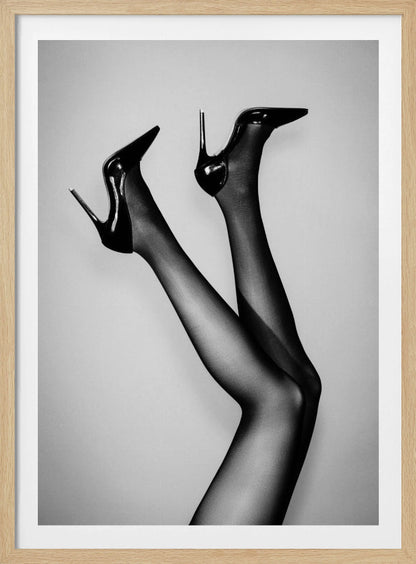 A black and white photograph of a woman's legs in the air, wearing sheer black stockings and shiny black stiletto high heels against a plain grey background. Poster