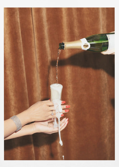 Champagne is poured from a bottle into a glass, overflowing with white foam that drips down onto two manicured hands with red nail polish holding it against a brown velvet curtain background. Poster