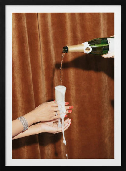 Champagne is poured from a bottle into a glass, overflowing with white foam that drips down onto two manicured hands with red nail polish holding it against a brown velvet curtain background. Poster