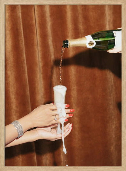Champagne is poured from a bottle into a glass, overflowing with white foam that drips down onto two manicured hands with red nail polish holding it against a brown velvet curtain background. Poster