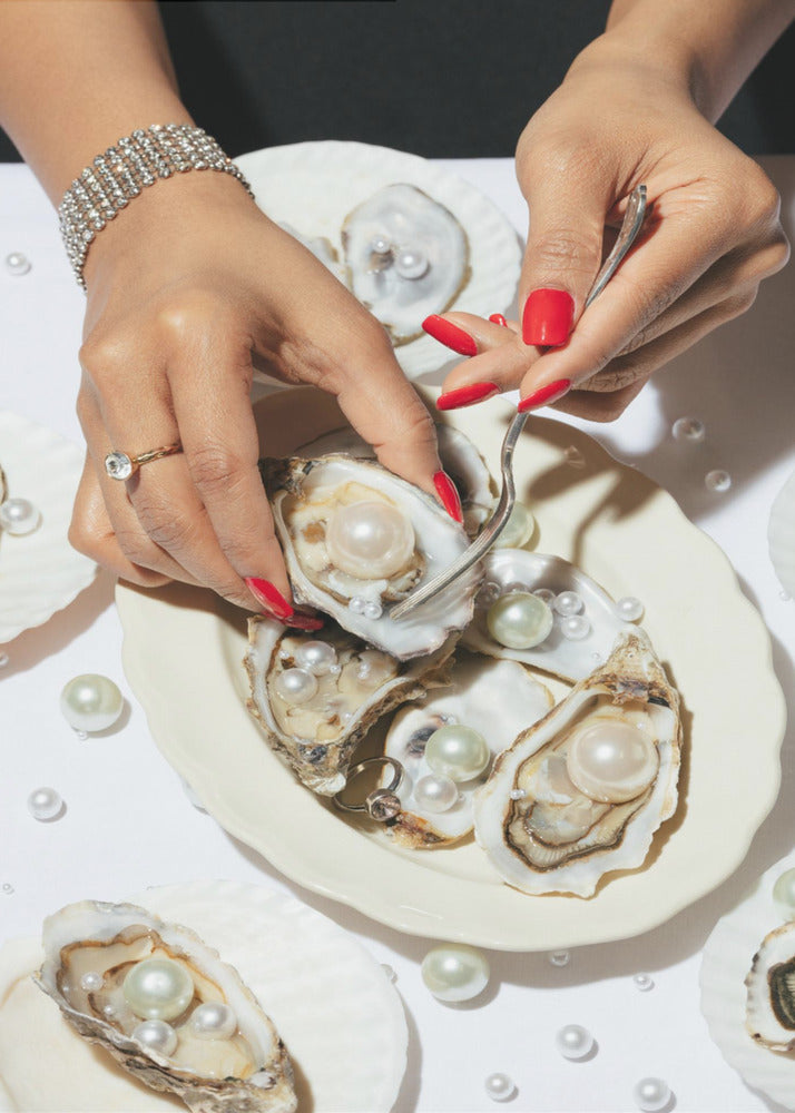 A close-up overhead shot of a woman's hands with vibrant red nails and elegant jewelry, using a fork to lift a large pearl from an oyster shell on a white plate. The plate and the surrounding white surface are adorned with numerous oysters filled with pearls of varying sizes, creating a luxurious and surreal scene. Print