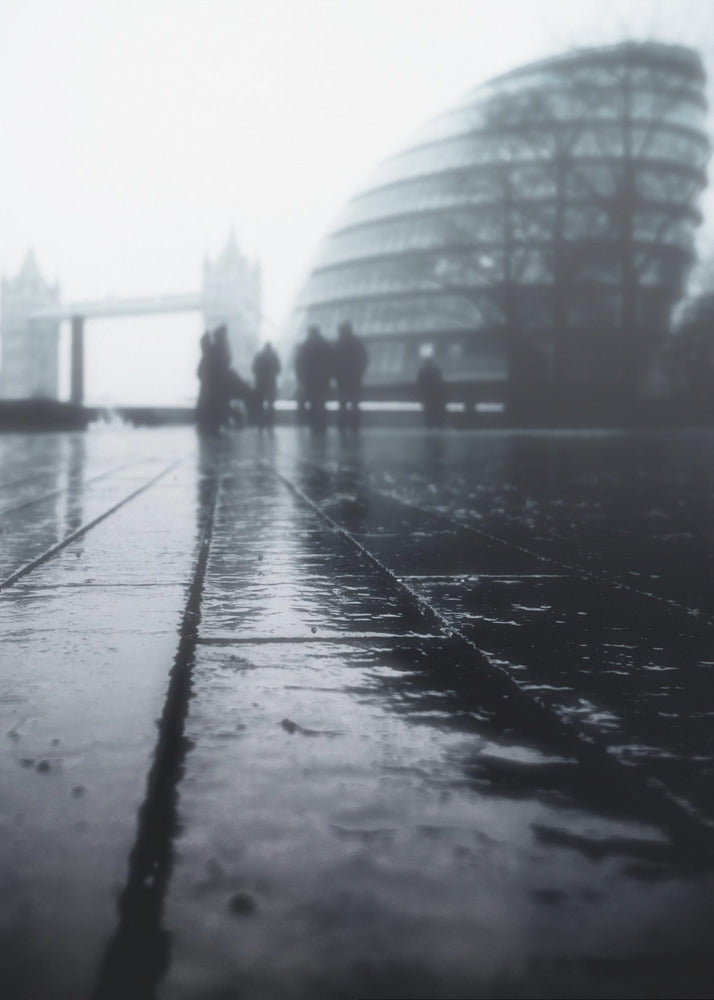 A moody, black and white, low-angle photograph of a wet city pavement in London. The foreground shows shimmering reflections on the dark, wet ground. In the blurry, foggy background, silhouettes of people stand near the modern City Hall building, with the iconic Tower Bridge visible further in the distance. Decor