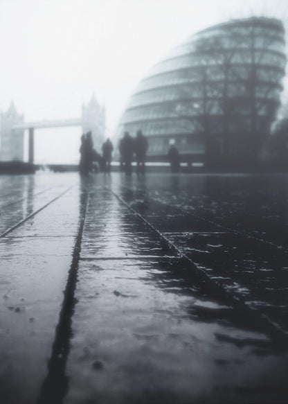 A moody, black and white, low-angle photograph of a wet city pavement in London. The foreground shows shimmering reflections on the dark, wet ground. In the blurry, foggy background, silhouettes of people stand near the modern City Hall building, with the iconic Tower Bridge visible further in the distance. Decor