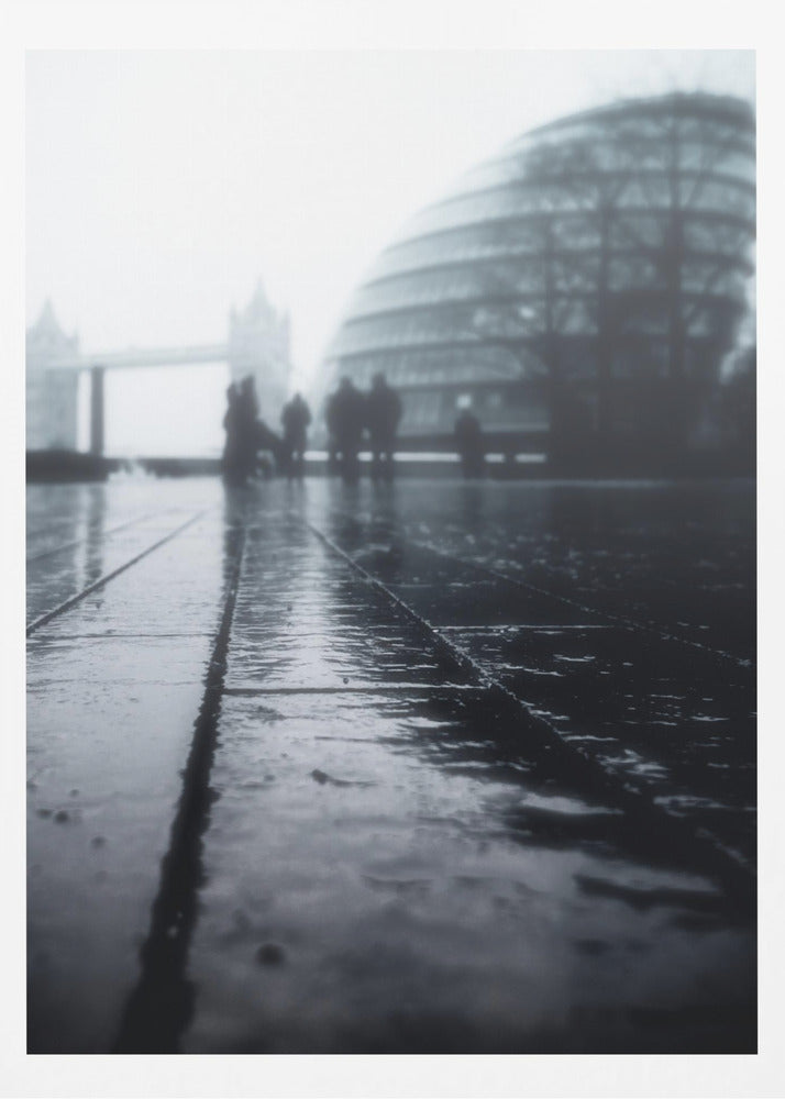 A moody, black and white, low-angle photograph of a wet city pavement in London. The foreground shows shimmering reflections on the dark, wet ground. In the blurry, foggy background, silhouettes of people stand near the modern City Hall building, with the iconic Tower Bridge visible further in the distance. Decor