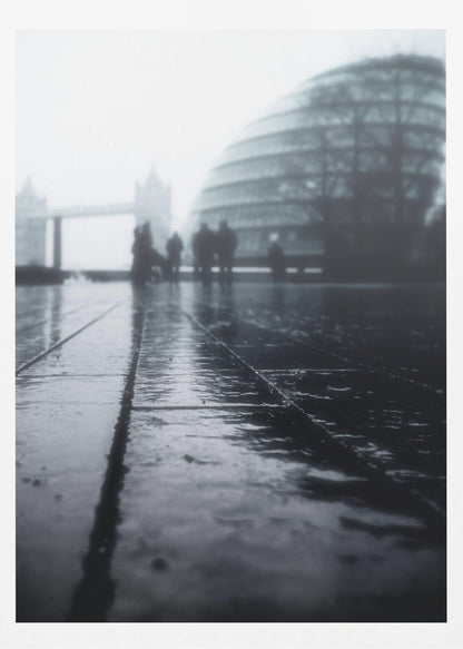 A moody, black and white, low-angle photograph of a wet city pavement in London. The foreground shows shimmering reflections on the dark, wet ground. In the blurry, foggy background, silhouettes of people stand near the modern City Hall building, with the iconic Tower Bridge visible further in the distance. Decor