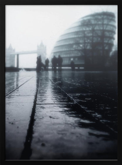 A moody, black and white, low-angle photograph of a wet city pavement in London. The foreground shows shimmering reflections on the dark, wet ground. In the blurry, foggy background, silhouettes of people stand near the modern City Hall building, with the iconic Tower Bridge visible further in the distance. Decor