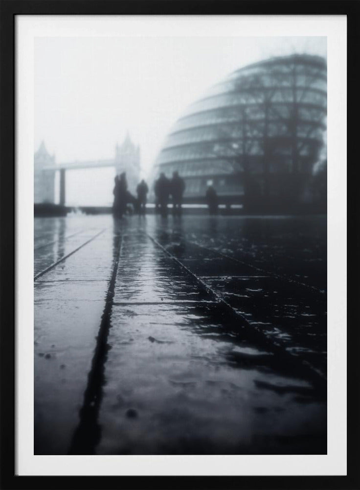 A moody, black and white, low-angle photograph of a wet city pavement in London. The foreground shows shimmering reflections on the dark, wet ground. In the blurry, foggy background, silhouettes of people stand near the modern City Hall building, with the iconic Tower Bridge visible further in the distance. Decor