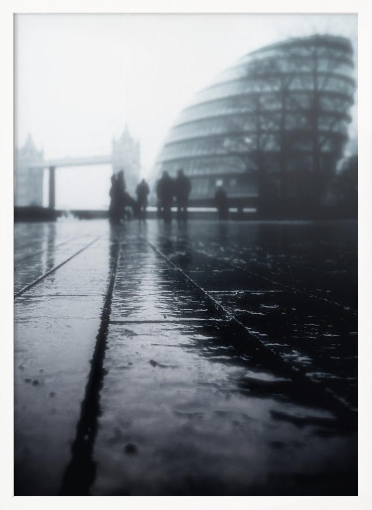 A moody, black and white, low-angle photograph of a wet city pavement in London. The foreground shows shimmering reflections on the dark, wet ground. In the blurry, foggy background, silhouettes of people stand near the modern City Hall building, with the iconic Tower Bridge visible further in the distance. Decor