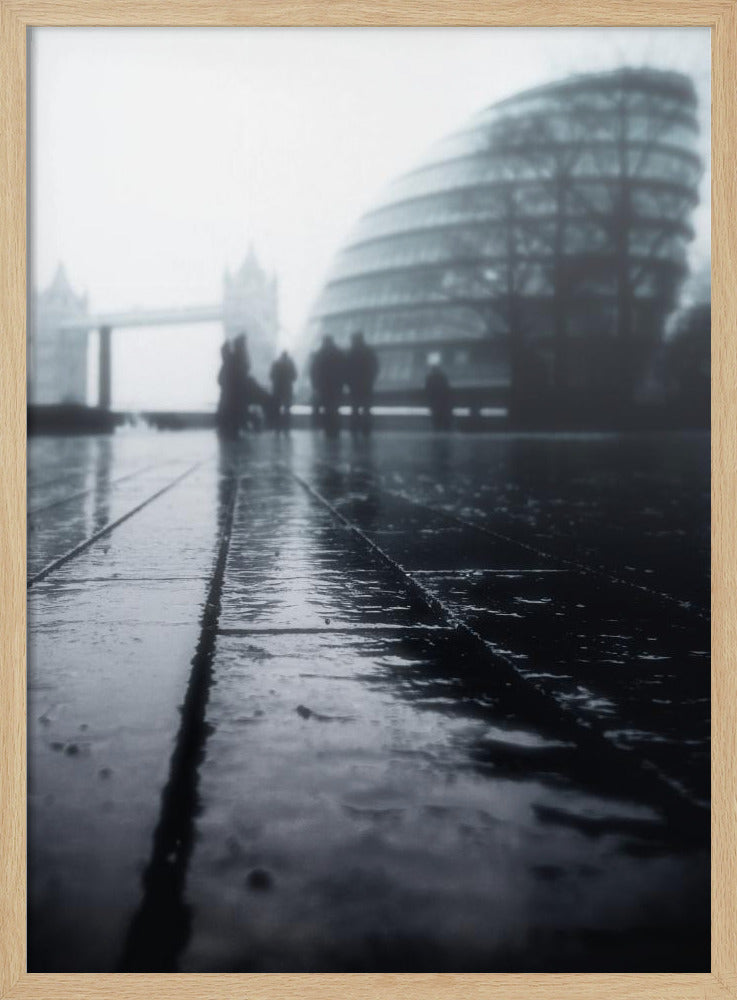 A moody, black and white, low-angle photograph of a wet city pavement in London. The foreground shows shimmering reflections on the dark, wet ground. In the blurry, foggy background, silhouettes of people stand near the modern City Hall building, with the iconic Tower Bridge visible further in the distance. Decor