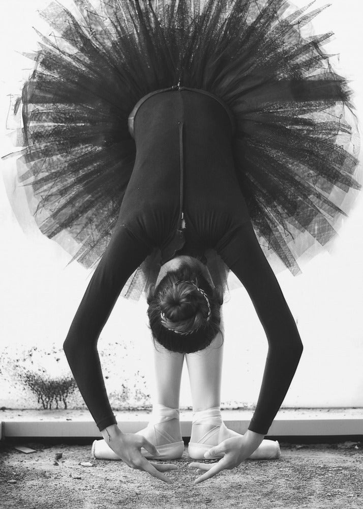 A striking black and white photograph of a ballerina in a deep forward bend. Her body is clad in a black leotard and white tights, with a voluminous black tutu fanning out vertically behind her. Her hair is tied in a bun, and her hands gracefully reach towards her feet, which are in white pointe shoes. Artwork
