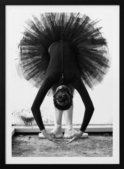 A striking black and white photograph of a ballerina in a deep forward bend. Her body is clad in a black leotard and white tights, with a voluminous black tutu fanning out vertically behind her. Her hair is tied in a bun, and her hands gracefully reach towards her feet, which are in white pointe shoes. Artwork