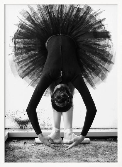 A striking black and white photograph of a ballerina in a deep forward bend. Her body is clad in a black leotard and white tights, with a voluminous black tutu fanning out vertically behind her. Her hair is tied in a bun, and her hands gracefully reach towards her feet, which are in white pointe shoes. Artwork