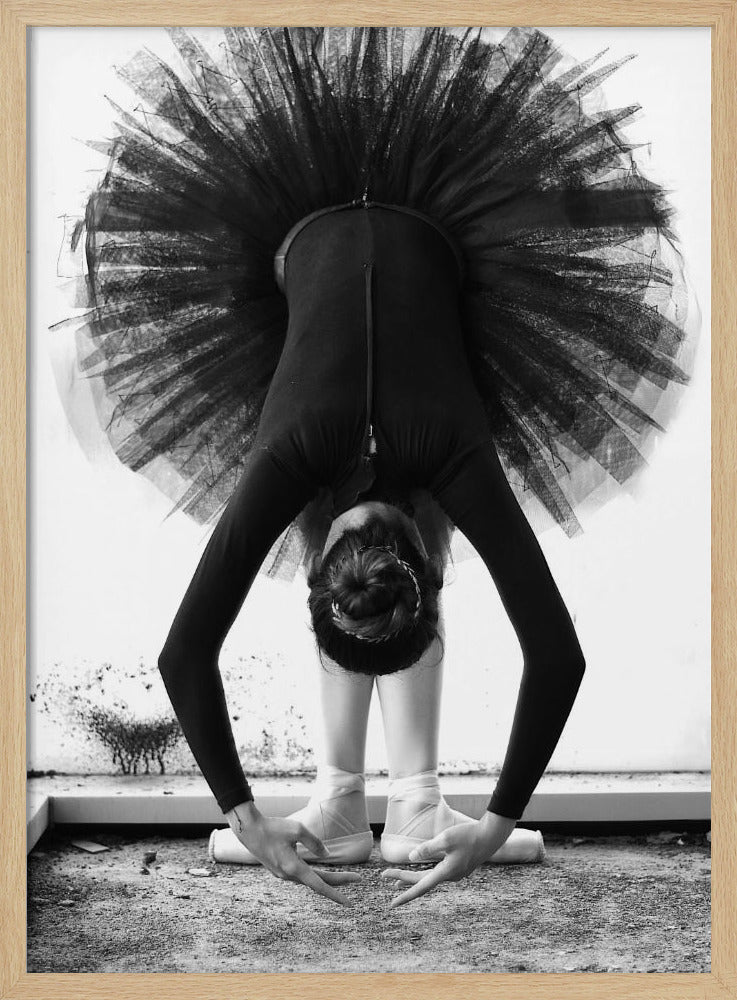 A striking black and white photograph of a ballerina in a deep forward bend. Her body is clad in a black leotard and white tights, with a voluminous black tutu fanning out vertically behind her. Her hair is tied in a bun, and her hands gracefully reach towards her feet, which are in white pointe shoes. Artwork