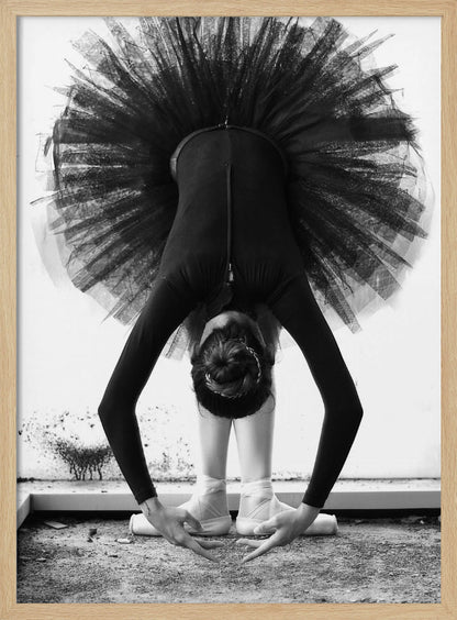 A striking black and white photograph of a ballerina in a deep forward bend. Her body is clad in a black leotard and white tights, with a voluminous black tutu fanning out vertically behind her. Her hair is tied in a bun, and her hands gracefully reach towards her feet, which are in white pointe shoes. Artwork