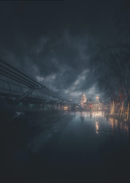 A moody, atmospheric night photograph of London, featuring the Millennium Bridge stretching across a wet surface towards the illuminated St. Paul's Cathedral under a dark, stormy sky. City lights reflect on the wet ground, and trees with warm lights line the right side of the frame. Artwork