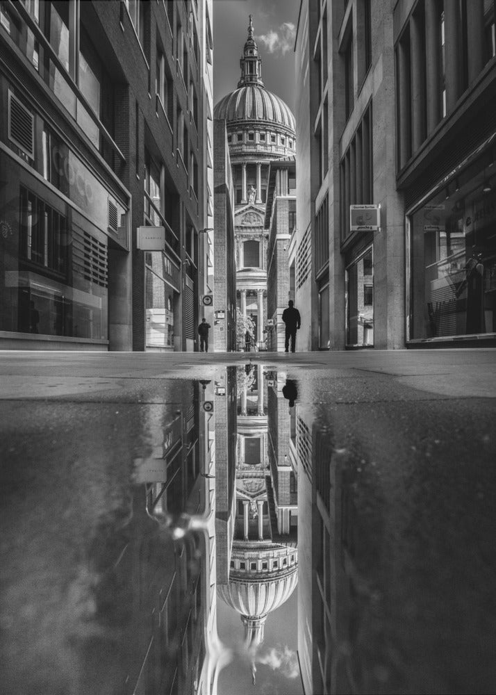 A dramatic black and white photograph taken from a low angle on a narrow city street, perfectly framing St. Paul's Cathedral in the distance. The foreground is dominated by a large puddle, which creates a stunning, clear reflection of the entire scene, including the cathedral, buildings, and sky. Print