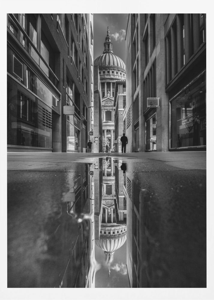 A dramatic black and white photograph taken from a low angle on a narrow city street, perfectly framing St. Paul's Cathedral in the distance. The foreground is dominated by a large puddle, which creates a stunning, clear reflection of the entire scene, including the cathedral, buildings, and sky. Print
