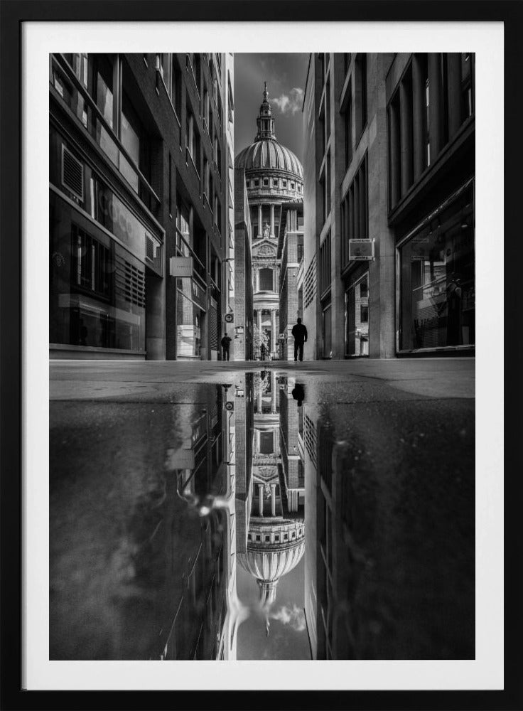 A dramatic black and white photograph taken from a low angle on a narrow city street, perfectly framing St. Paul's Cathedral in the distance. The foreground is dominated by a large puddle, which creates a stunning, clear reflection of the entire scene, including the cathedral, buildings, and sky. Print