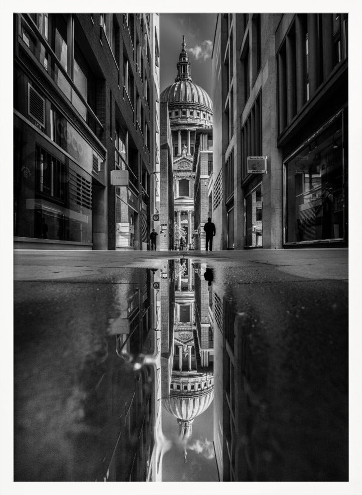 A dramatic black and white photograph taken from a low angle on a narrow city street, perfectly framing St. Paul's Cathedral in the distance. The foreground is dominated by a large puddle, which creates a stunning, clear reflection of the entire scene, including the cathedral, buildings, and sky. Print