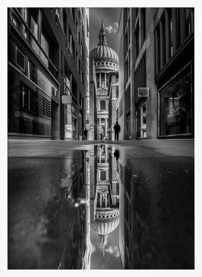 A dramatic black and white photograph taken from a low angle on a narrow city street, perfectly framing St. Paul's Cathedral in the distance. The foreground is dominated by a large puddle, which creates a stunning, clear reflection of the entire scene, including the cathedral, buildings, and sky. Print