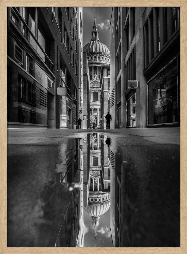 A dramatic black and white photograph taken from a low angle on a narrow city street, perfectly framing St. Paul's Cathedral in the distance. The foreground is dominated by a large puddle, which creates a stunning, clear reflection of the entire scene, including the cathedral, buildings, and sky. Print