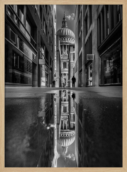 A dramatic black and white photograph taken from a low angle on a narrow city street, perfectly framing St. Paul's Cathedral in the distance. The foreground is dominated by a large puddle, which creates a stunning, clear reflection of the entire scene, including the cathedral, buildings, and sky. Print