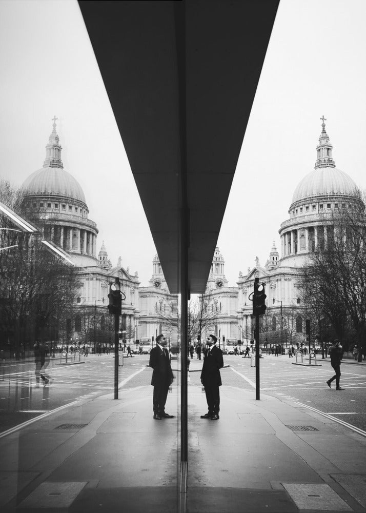 A surreal black and white photograph capturing a symmetrical reflection of a city scene, with St. Paul's Cathedral prominently featured on both sides. A dark, V-shaped reflection from a building divides the image vertically. In the foreground, the reflection of two men in dark suits standing on the sidewalk adds a human element to the grand architectural view. Decor