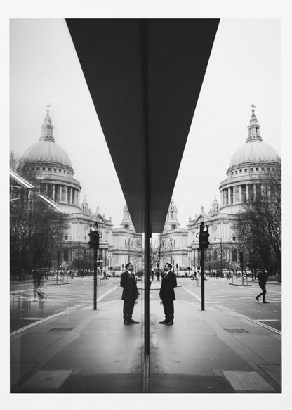 A surreal black and white photograph capturing a symmetrical reflection of a city scene, with St. Paul's Cathedral prominently featured on both sides. A dark, V-shaped reflection from a building divides the image vertically. In the foreground, the reflection of two men in dark suits standing on the sidewalk adds a human element to the grand architectural view. Decor