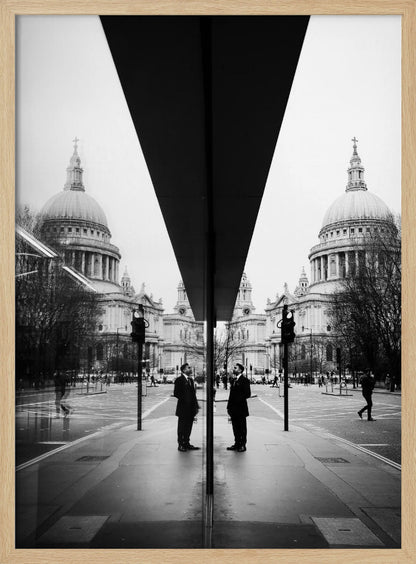 A surreal black and white photograph capturing a symmetrical reflection of a city scene, with St. Paul's Cathedral prominently featured on both sides. A dark, V-shaped reflection from a building divides the image vertically. In the foreground, the reflection of two men in dark suits standing on the sidewalk adds a human element to the grand architectural view. Decor