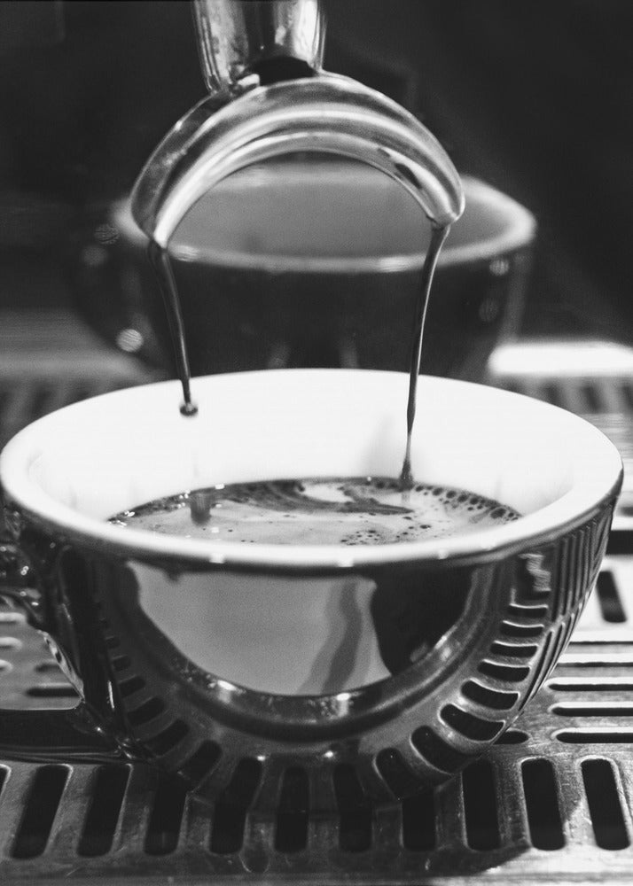 A close-up, black and white artistic photograph capturing the moment two streams of dark espresso pour from a portafilter into a waiting cup on a coffee machine's drip tray. Wall Art
