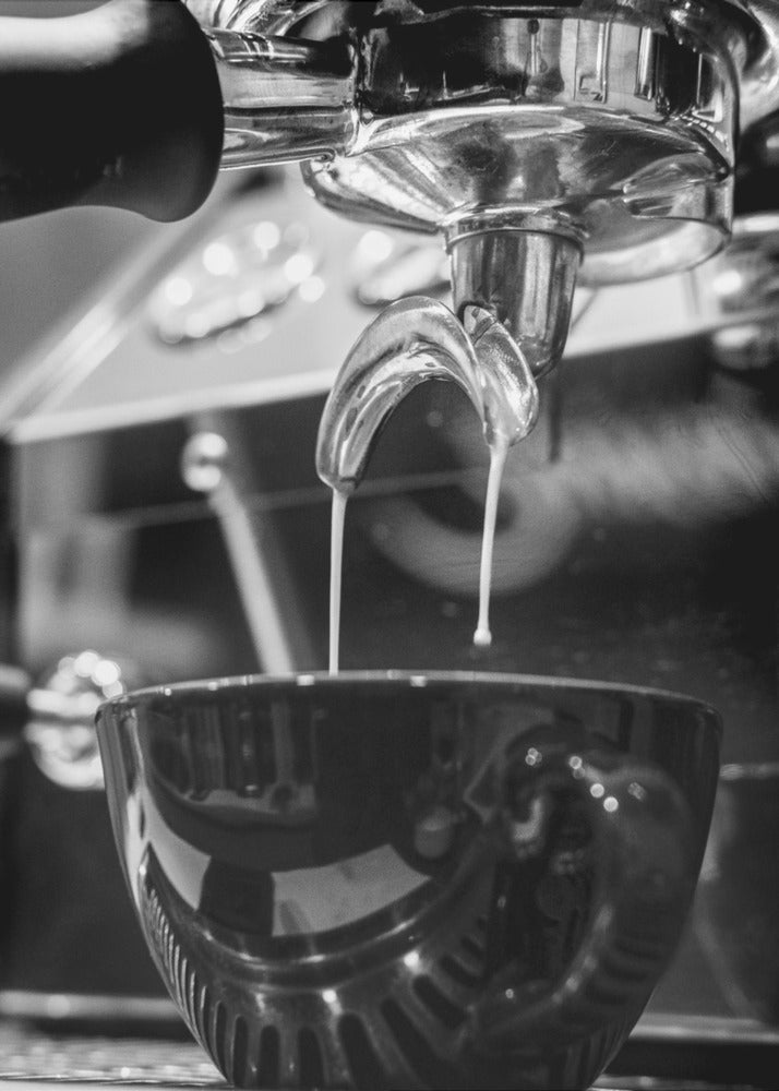 A close-up, black and white photograph of an espresso machine pouring two streams of fresh coffee from a chrome portafilter into a dark, glossy cup. Print