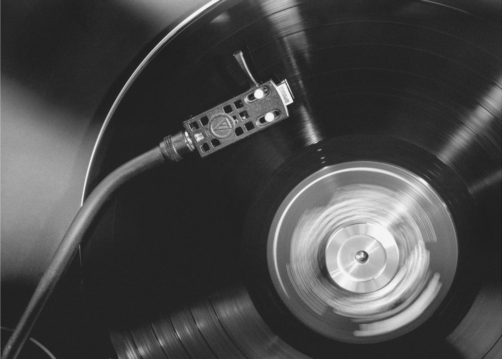 A framed, black and white, close-up photograph of a record player in action. The turntable is spinning a vinyl record, creating a motion blur on the center label. The tonearm and stylus are positioned over the grooved surface of the record. Poster