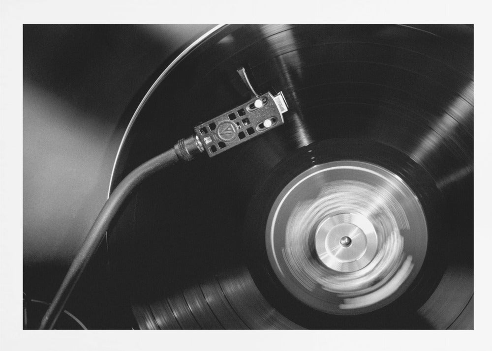 A framed, black and white, close-up photograph of a record player in action. The turntable is spinning a vinyl record, creating a motion blur on the center label. The tonearm and stylus are positioned over the grooved surface of the record. Poster