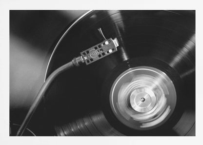 A framed, black and white, close-up photograph of a record player in action. The turntable is spinning a vinyl record, creating a motion blur on the center label. The tonearm and stylus are positioned over the grooved surface of the record. Poster