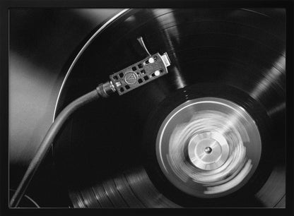 A framed, black and white, close-up photograph of a record player in action. The turntable is spinning a vinyl record, creating a motion blur on the center label. The tonearm and stylus are positioned over the grooved surface of the record. Poster
