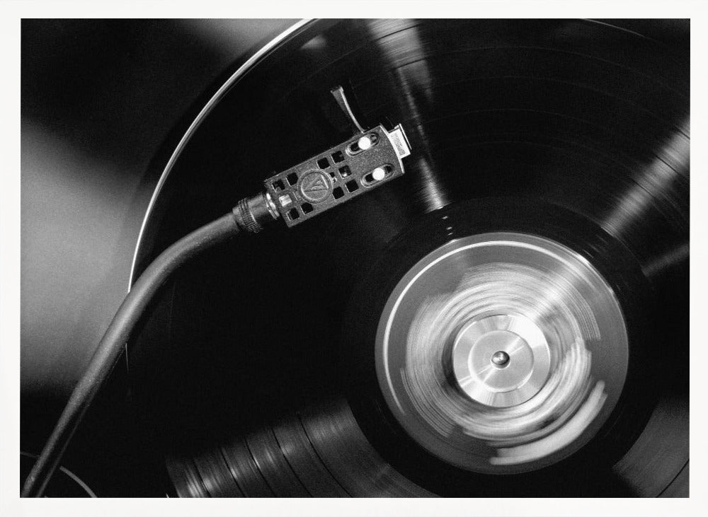 A framed, black and white, close-up photograph of a record player in action. The turntable is spinning a vinyl record, creating a motion blur on the center label. The tonearm and stylus are positioned over the grooved surface of the record. Poster