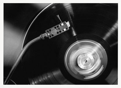 A framed, black and white, close-up photograph of a record player in action. The turntable is spinning a vinyl record, creating a motion blur on the center label. The tonearm and stylus are positioned over the grooved surface of the record. Poster