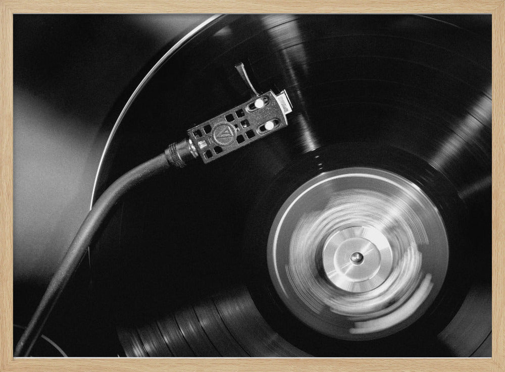 A framed, black and white, close-up photograph of a record player in action. The turntable is spinning a vinyl record, creating a motion blur on the center label. The tonearm and stylus are positioned over the grooved surface of the record. Poster