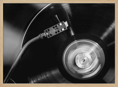 A framed, black and white, close-up photograph of a record player in action. The turntable is spinning a vinyl record, creating a motion blur on the center label. The tonearm and stylus are positioned over the grooved surface of the record. Poster