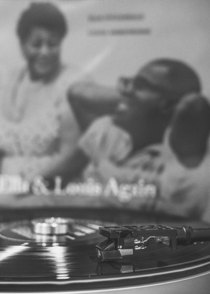 A black and white photograph focusing on a turntable's stylus resting on a spinning vinyl record. In the blurred background, the album cover for 'Ella & Louis Again' featuring Ella Fitzgerald and Louis Armstrong is visible. Poster
