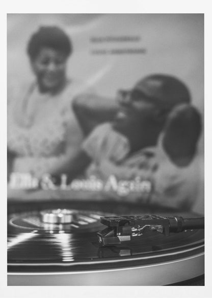 A black and white photograph focusing on a turntable's stylus resting on a spinning vinyl record. In the blurred background, the album cover for 'Ella & Louis Again' featuring Ella Fitzgerald and Louis Armstrong is visible. Poster