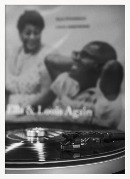 A black and white photograph focusing on a turntable's stylus resting on a spinning vinyl record. In the blurred background, the album cover for 'Ella & Louis Again' featuring Ella Fitzgerald and Louis Armstrong is visible. Poster