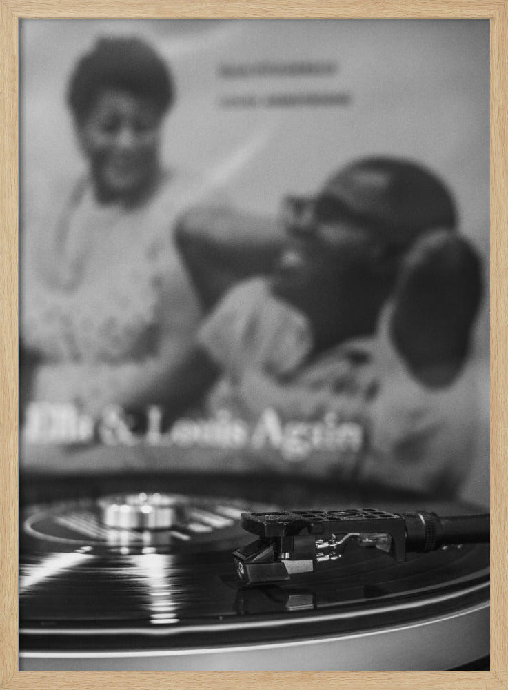 A black and white photograph focusing on a turntable's stylus resting on a spinning vinyl record. In the blurred background, the album cover for 'Ella & Louis Again' featuring Ella Fitzgerald and Louis Armstrong is visible. Poster