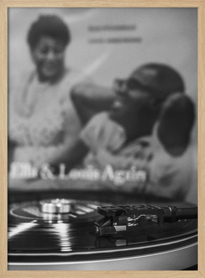 A black and white photograph focusing on a turntable's stylus resting on a spinning vinyl record. In the blurred background, the album cover for 'Ella & Louis Again' featuring Ella Fitzgerald and Louis Armstrong is visible. Poster
