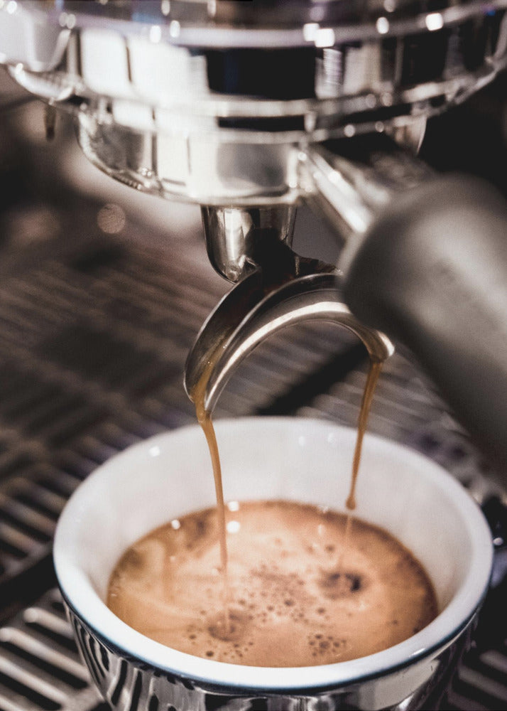 A close-up photograph of a shiny chrome espresso machine brewing a fresh shot of espresso. Two streams of rich, dark coffee pour from the portafilter into a white ceramic cup with a blue patterned rim, creating a light brown crema on top. Print
