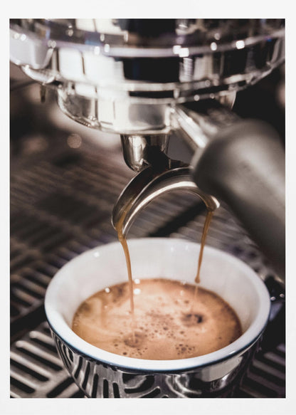 A close-up photograph of a shiny chrome espresso machine brewing a fresh shot of espresso. Two streams of rich, dark coffee pour from the portafilter into a white ceramic cup with a blue patterned rim, creating a light brown crema on top. Print