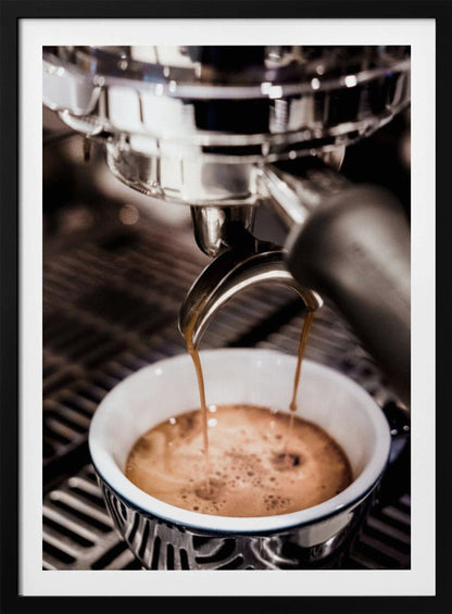 A close-up photograph of a shiny chrome espresso machine brewing a fresh shot of espresso. Two streams of rich, dark coffee pour from the portafilter into a white ceramic cup with a blue patterned rim, creating a light brown crema on top. Print