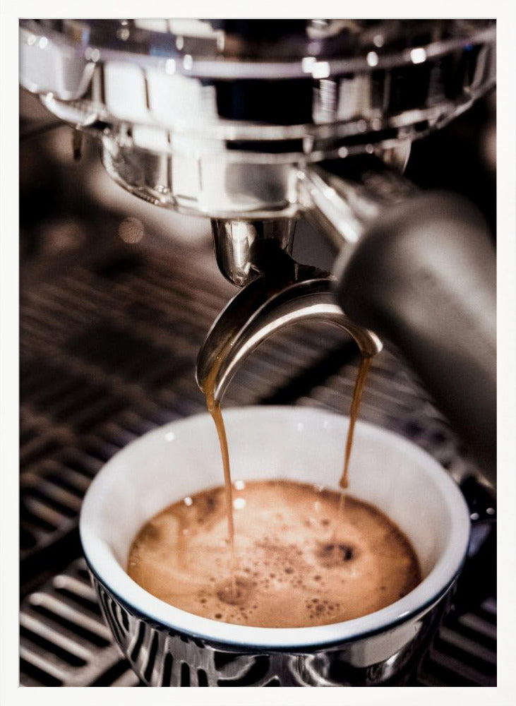 A close-up photograph of a shiny chrome espresso machine brewing a fresh shot of espresso. Two streams of rich, dark coffee pour from the portafilter into a white ceramic cup with a blue patterned rim, creating a light brown crema on top. Print