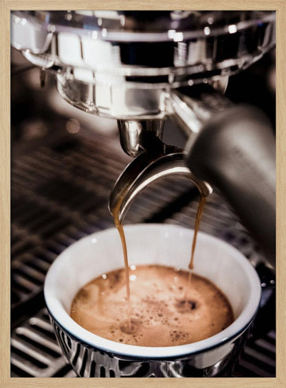 A close-up photograph of a shiny chrome espresso machine brewing a fresh shot of espresso. Two streams of rich, dark coffee pour from the portafilter into a white ceramic cup with a blue patterned rim, creating a light brown crema on top. Print